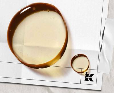 A close-up image of two circular droplets of a brown liquid on a white background, with one droplet larger than the other, positioned next to a black letter "K" in the corner.