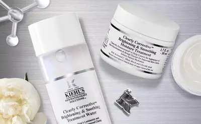 Alt text: Two skincare products from Kiehl's: a bottle of Clearly Corrective Brightening & Soothing Treatment Water and a jar of Clearly Corrective Brightening & Smoothing Moisture Treatment, placed on a textured surface with a white flower and a small pin in the background.