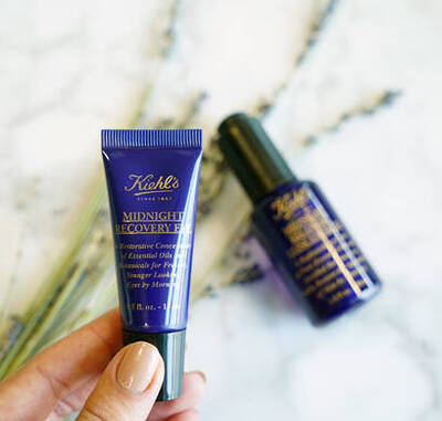 Una mano sostiene un tubo de crema Kiehl's Midnight Recovery junto a un envase negro de aceite, con flores secas al fondo.