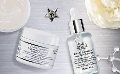 A flat lay of skincare products on a silver background, featuring a jar labeled "Brightening & Smoothing Moisture Treatment" and a dropper bottle labeled "Dark Spot Solution," both by Kiehl's. A star emblem is in the top left corner.
