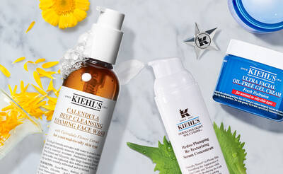 A flat lay of Kiehl's skincare products on a marble surface, including a foaming face wash, a hydrating serum, and an oil-free gel cream, surrounded by yellow flower petals and green leaves.