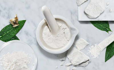A white mortar and pestle surrounded by scattered white powder, leaves, and a small dish with more powder on a marble surface.