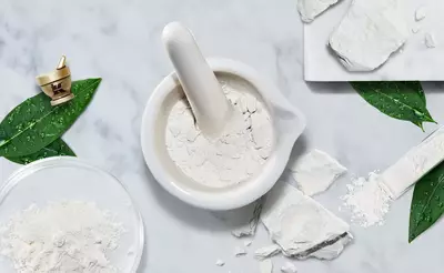 A white mortar and pestle surrounded by scattered white powder, leaves, and a small dish with more powder on a marble surface.