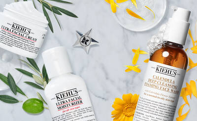 A flat lay of Kiehl's skincare products on a marble surface, featuring Ultra Facial Cream, Ultra Facial Moisturizer, and Calendula Deep Cleansing Foaming Face Wash, surrounded by yellow flowers and green leaves.