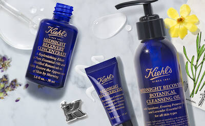 A collection of Kiehl's Midnight Recovery skincare products featuring a blue glass bottle of concentrate, a tube of eye cream, and a pump bottle of cleansing oil, styled with floral elements and a small letter K charm.