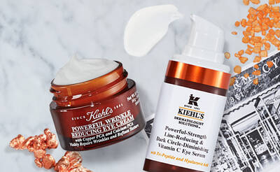 Alt text: A flat lay image of Kiehl's skincare products, including a jar of Powerful Wrinkle Reducing Eye Cream and a pump bottle of Powerful-Strength Dark Circle-Diminishing Vitamin C Eye Serum, surrounded by orange granules.