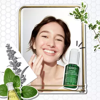 Mujer sonriente aplicando serum de aceite de cannabis de Kiehl's frente a un espejo. Elementos botanicos alrededor.