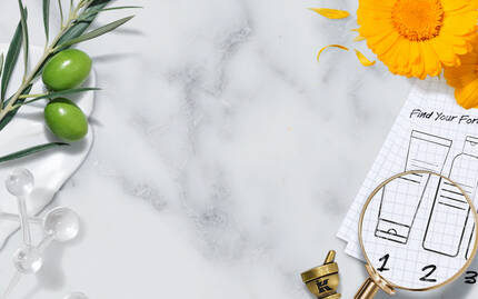 A flat lay featuring a marble background with various natural ingredients, including olive branches and herbal elements, alongside a magnifying glass and a blueprint of product formulas. A blank white area in the center is framed by an elegant border.