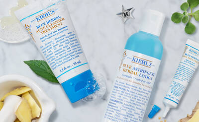 A flat lay of Kiehl's skincare products, including a blue herbal acne treatment, blue astringent herbal lotion, and a small tube, surrounded by herbs and natural ingredients.