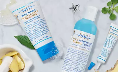 A flat lay of Kiehl's skincare products, including a blue herbal acne treatment, blue astringent herbal lotion, and a small tube, surrounded by herbs and natural ingredients.