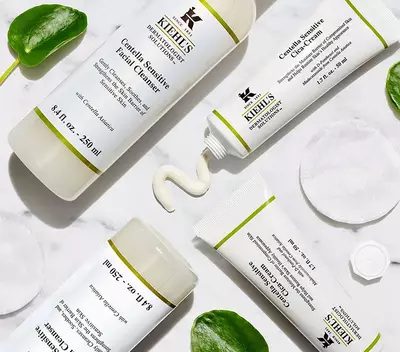 Productos de Kiehl's: limpiador facial y crema, sobre una superficie de marmol con discos de algodon y hojas decorativas.