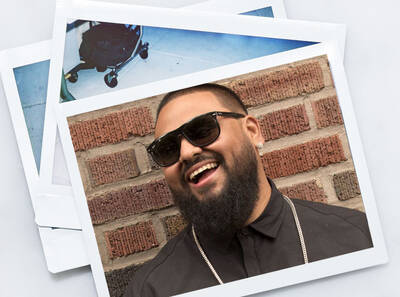 Hombre con barba, gafas de sol y camiseta negra sonrie frente a una pared de ladrillos, con fotos en el fondo.