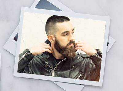 Hombre con barba y cabello corto, vestido con chaqueta de cuero, posando de forma relajada frente a una pared.