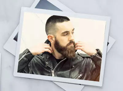 Hombre con barba y cabello corto, vestido con chaqueta de cuero, posando de forma relajada frente a una pared.
