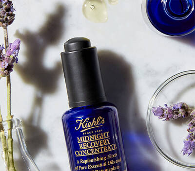 Botella de suero nocturno de Kiehl's sobre una superficie blanca, rodeada de lavanda y gotas de aceite.