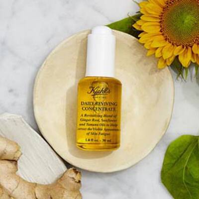 Frasco de serum facial Kiehl's Daily Reviving Concentrate sobre un plato, con jengibre, girasol y hojas verdes de fondo.