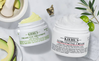 Two jars of Kiehl's skincare products are displayed on a marble surface. One jar is labeled "Creamy Eye Treatment with Avocado" and the other is "Ultra Facial Cream." There are sliced avocados and a small mortar in the background, enhancing the natural theme.