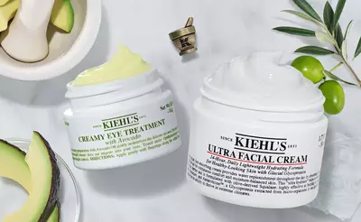 Two jars of Kiehl's skincare products are displayed on a marble surface. On the left is the "Creamy Eye Treatment with Avocado," and on the right is the "Ultra Facial Cream." In the background, there are avocado slices and a small mortar and pestle.