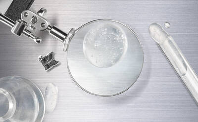 A close-up image featuring a magnifying glass, a dropper, and a glass vial on a metallic surface, with a droplet of liquid reflecting light.