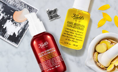 An arrangement of Kiehl's skincare products featuring a red Glow Formula Skin Hydrator and a yellow Daily Reviving Concentrate, surrounded by ginger root slices and flower petals, with a vintage pharmacy photo in the background.
