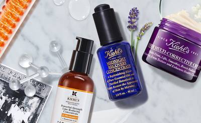 A flat lay of Kiehl's skincare products, including a brown bottle of Powerful-Strength Line-Reducing Concentrate, a blue bottle of Midnight Recovery Concentrate, and a purple jar of Super Multi-Corrective Cream, surrounded by various skincare-related elements.