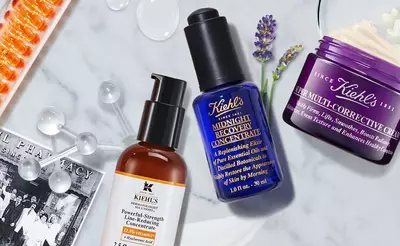 A flat lay of Kiehl's skincare products, including a brown bottle of Powerful-Strength Line-Reducing Concentrate, a blue bottle of Midnight Recovery Concentrate, and a purple jar of Super Multi-Corrective Cream, surrounded by various skincare-related elements.
