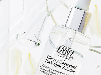 A bottle of Kiehl's Clearly Corrective Dark Spot Solution surrounded by delicate flower petals and a glass container.