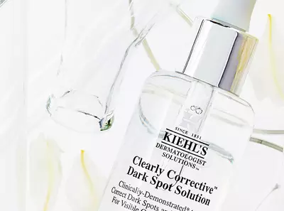 A bottle of Kiehl's Clearly Corrective Dark Spot Solution surrounded by delicate flower petals and a glass container.