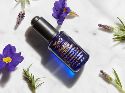 Frasco de suero hidratante de Kiehl's sobre una superficie blanca, rodeado de flores moradas y hierbas.