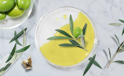 A glass bowl filled with bright yellow olive oil, topped with a green olive and olive leaves, surrounded by a plate of green olives and scattered olive leaves on a marble surface.