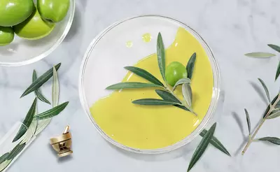 A glass bowl filled with bright yellow olive oil, topped with a green olive and olive leaves, surrounded by a plate of green olives and scattered olive leaves on a marble surface.
