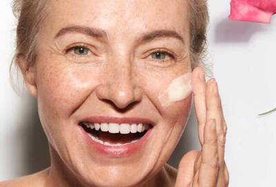 Mujer sonriente aplicandose crema en la cara, con un fondo de rosas y luz suave.