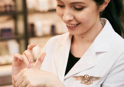 Mujer sonriendo mientras aplica un producto en la mano, vistiendo una bata con el logo de Kiehl's.