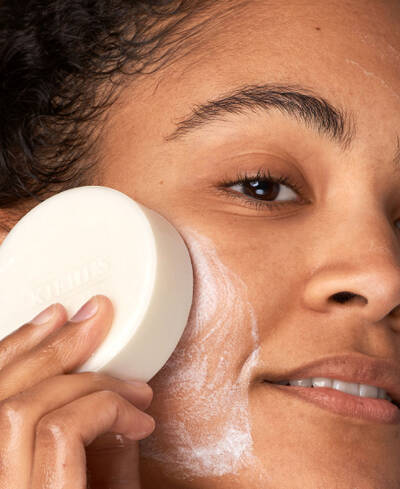 Mujer aplicando un jabon facial en su piel, sonriendo mientras limpia su rostro con suavidad.