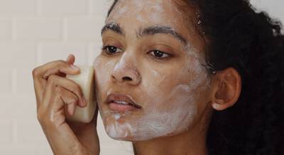 Mujer aplicando jabon facial espumoso en su rostro mientras se mira en un espejo.