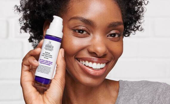 Una mujer sonriente con cabello rizado sostiene un suero Kiehl's
Retinol Daily
Micro-Dose cerca de su rostro.