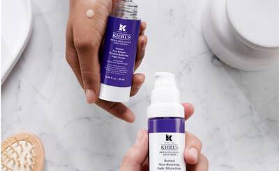 Manos sosteniendo productos de Kiehl's: un serum y una crema con envases morados, sobre una superficie clara y minimalista.