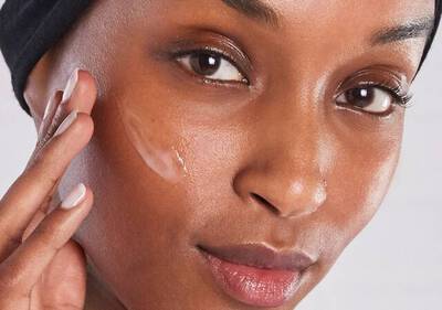 Mujer aplicando crema facial en su piel, sonriente, con un fondo suave que destaca su rostro y mirada natural.