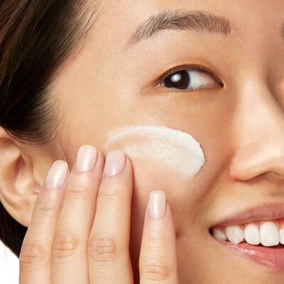 Mujer aplicando crema hidratante en su rostro, sonriendo y mostrando una piel radiante y cuidada.