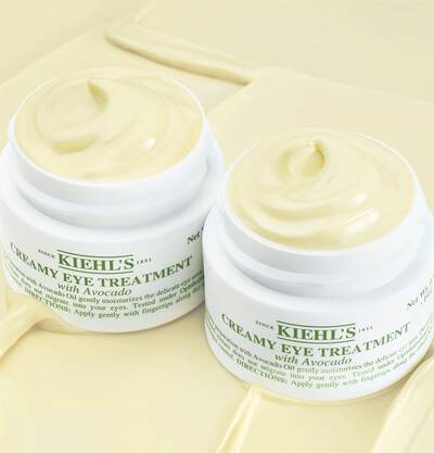 Tratamiento cremoso para ojos de Kiehl's con aguacate, presentado en frascos blancos sobre un fondo suave y cremoso.