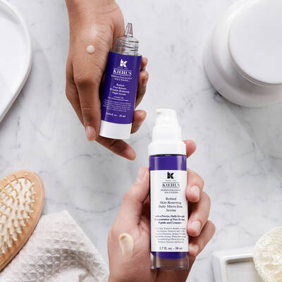Dos frascos de productos para la piel de Kiehl's, sostenidos por una mano, sobre una superficie de marmol.