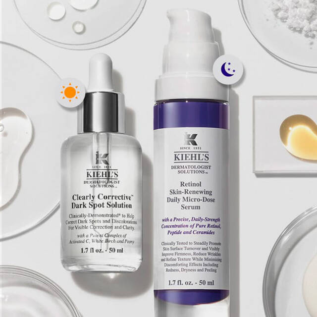 Imagen de productos de cuidado de la piel de Kiehl's dispuestos sobre una superficie blanca, incluyendo serums y tratamientos en frascos y goteros, acompanados de ingredientes naturales como rodajas de naranja y gotas.