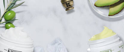 Imagen de dos envases de crema sobre una superficie de marmol. Rodeados de ingredientes naturales como aceitunas y aguacate.