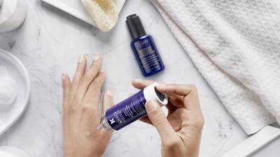 Unas manos aplicando un suero facial sobre una superficie de marmol con productos de cuidado de la piel en el fondo.