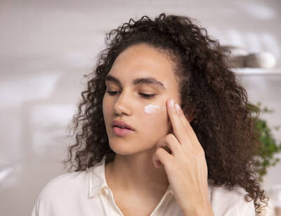 Mujer con cabello rizado aplica crema facial en su mejilla, en un entorno luminoso y moderno.