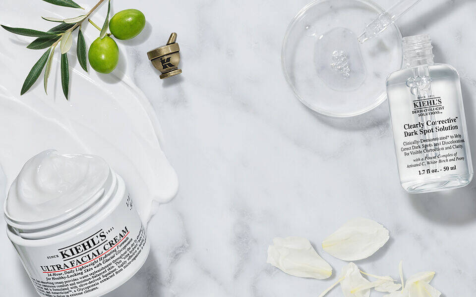 Imagen de productos de cuidado de la piel de Kiehl's, incluyendo una crema, un aceite corporal y una locion, dispuestos sobre una superficie de marmol con algunos ingredientes naturales.
